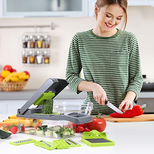 Happy woman in a green striped shirt in a kitchen using a vegetable chopper to slice a cucumber. On the counter are various vegetables and the chopper's multiple blade attachments are laid out