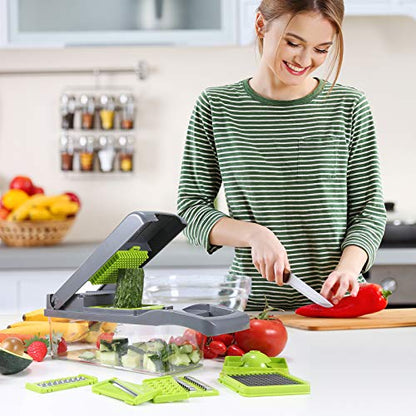 Happy woman in a green striped shirt in a kitchen using a vegetable chopper to slice a cucumber. On the counter are various vegetables and the chopper's multiple blade attachments are laid out